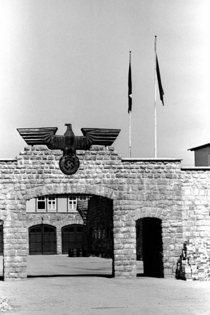 Steintor gekrönt mit einem großen Metalladler, der ein Hakenkreuz hält; durch das Tor ist ein Gebäude mit zwei Garagentoren sichtbar