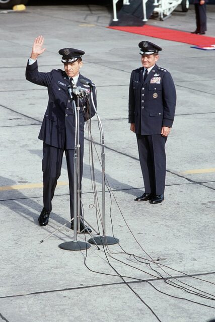 James Risner speaking at a microphone, with an unidentified serviceman standing near him