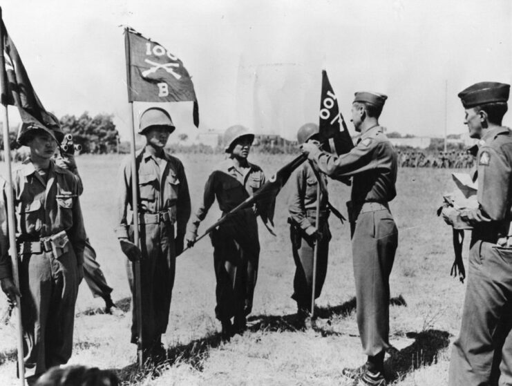 Mark Clark fastening citation streamers on the quidons of the 100th Infantry Battalion's flag, while soldiers holding flags stand before him