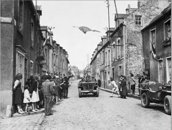 The French Tricolour floats over a street in Carentan, the first French town to be liberated by the Americans. French civilians wave their hands in welcome as men of the US 7th Corps pass through the streets after two days of bitter fighting.