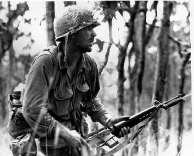 Rick Rescorla holding a rifle in the middle of a wooded area