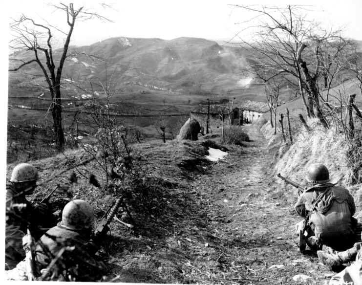 Members of Company K, 87th Mountain Infantry Regiment, 10th Mountain Division crouching on the edge of a dirt road