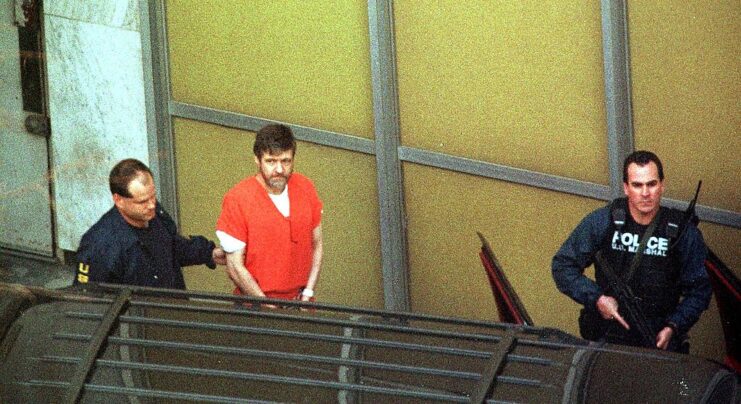Two police officers leading Ted Kaczynski, in handcuffs, along the side of a building
