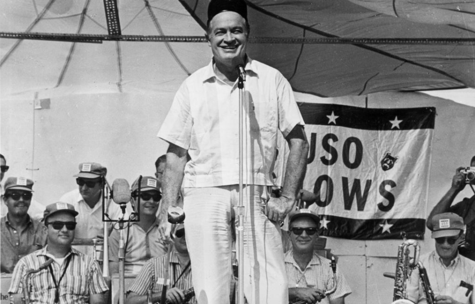 Bob Hope performing on stage, with a USO flag hanging behind him