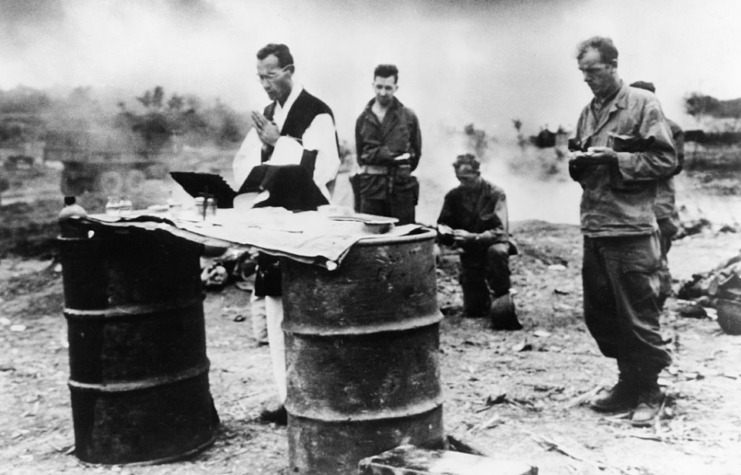 Clergyman praying while American servicemen watch from behind