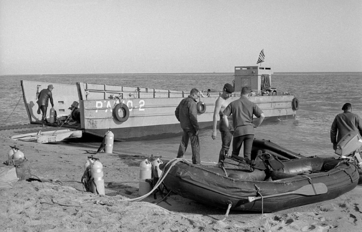 US Navy frogmen standing next to a boat and inflatable raft on the beach