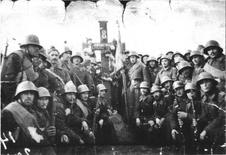 Black and white, group of men in military uniforms and waring helmets stand and kneel in front of a monument shaped like a cross. 