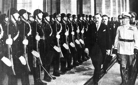 Black and white, man in suit and man in white military uniform walk past soldiers standing at attention in black uniforms holding guns and wearing helmets. 