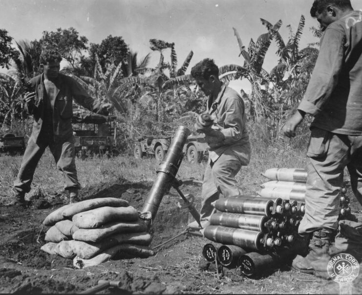 Black and white. Two men stand on either side of a man kneeling loading a 4.2 inch mortar outside on grass.