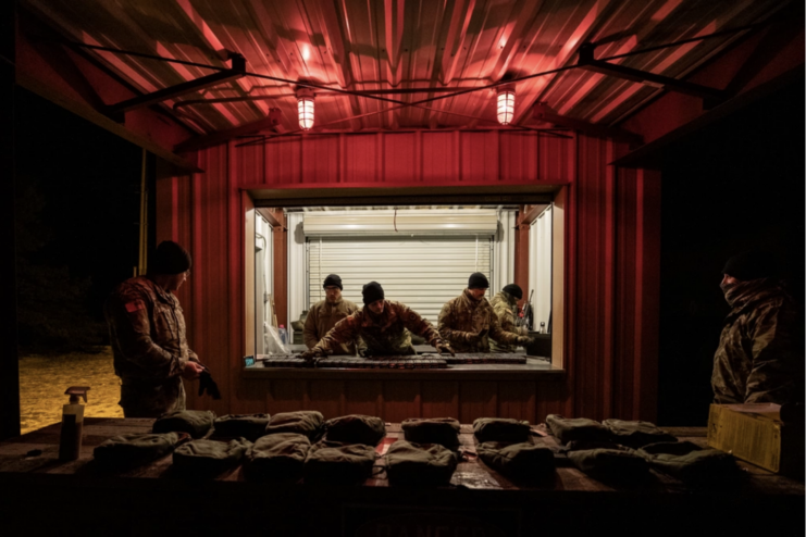 Dark room with two red lights on the ceiling. Ammo and gear on the table in the middle of the room. two men in uniform stand on either side of the table and four men stand in a room with the lights on in the middle of the picture and look through a window into the dark room. 