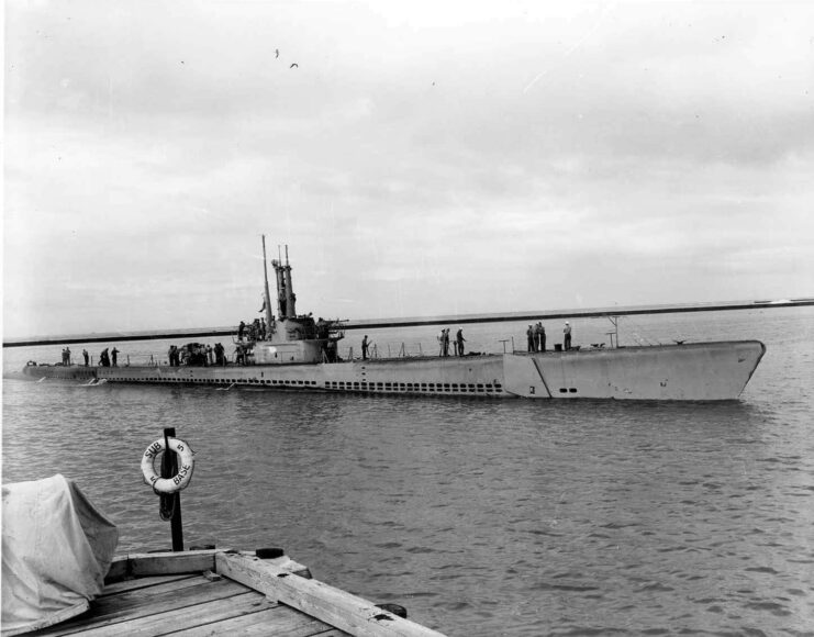 Black and white. Top of submarine shown above water's surface. corner of a dock can be seen in the bottom left corner.