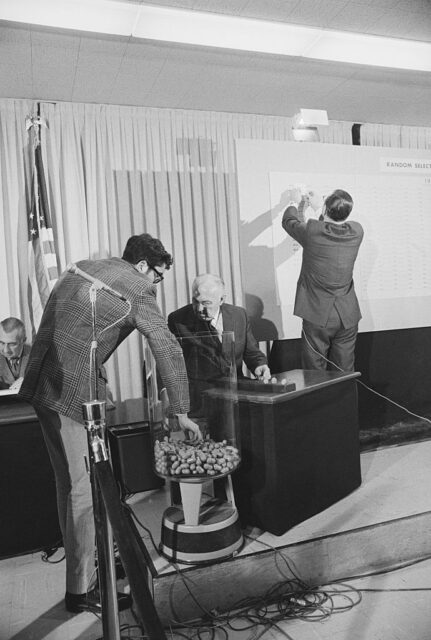 Paul M. Murray grabbing a capsule from a jar while Daniel Omer sits at a desk and Charles Fox is turned toward a large board