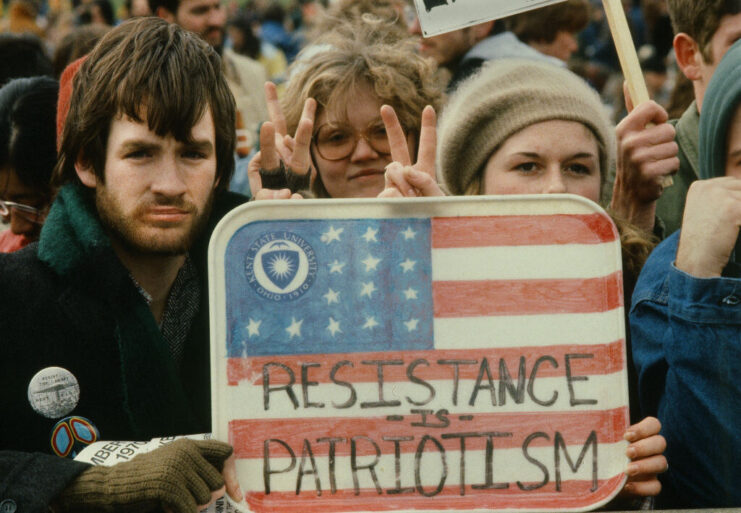 Protestors holding a sign with the American flag and "RESISTANCE IS PATRIOTISM" painted on it