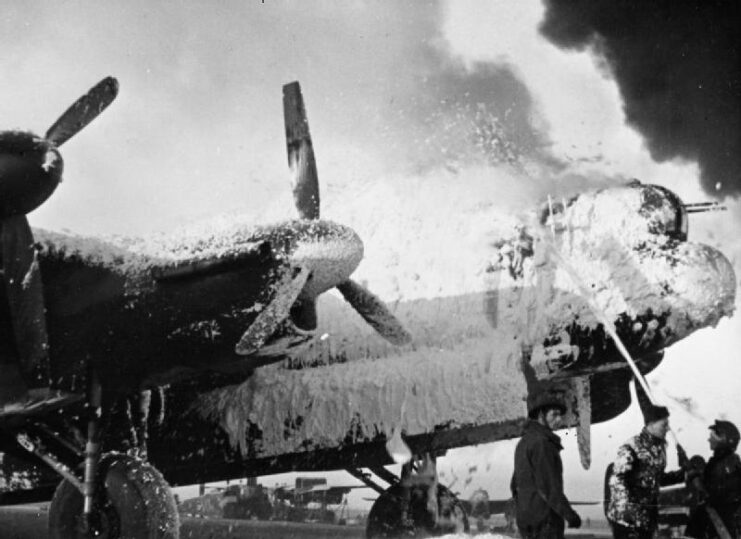 Black and white. Up-angle shot of the front of a bomber with foam being sprayed on it and covering the front. Smoke seen in the background. 3 men holding the hose that is spraying the foam. 