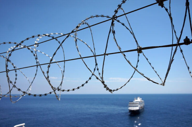 Barbed wire with water and blue sky and a boat in the background. 