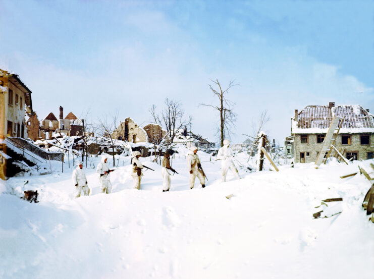 6 men in white military uniforms and holding guns stand on top of a snowbank in a line. Blue sky behind them and one house can be seen on either side of the men with two behind them in the distance. 