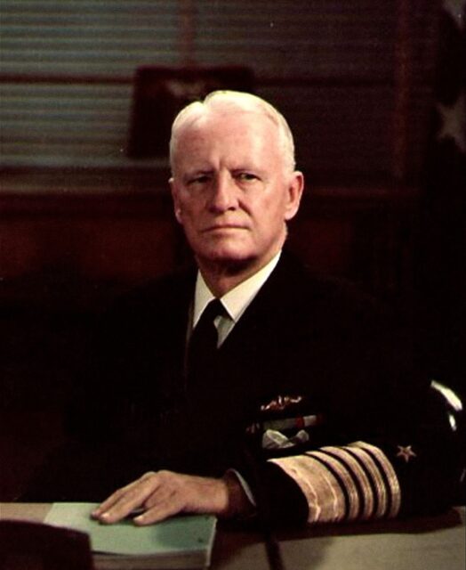 Man with white hair sitting behind a desk while wearing a naval uniform. One hand resting on the desk. 