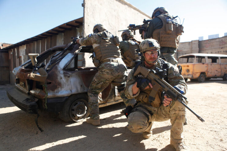 Chris Kyle holding a rifle kneeling in front of men pointing guns over a destroyed car behind him.