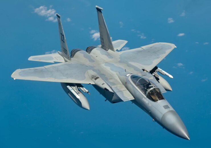 Top right side of gray camo colored F-15C Eagle flying in blue sky with some small white clouds. 