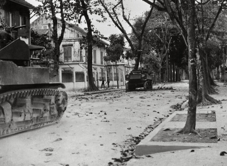 Two tanks moving along a street, past two soldiers