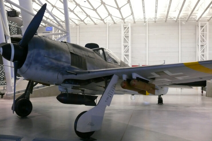 Front left side of Focke Wulf FW190F-8 in room with white floors and walls and white scaffolding ceiling. 