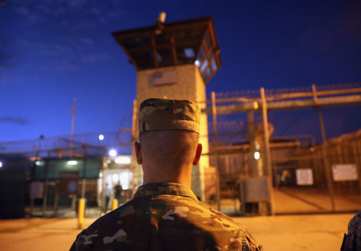 Back of man's head and shoulders, dressed in a camo uniform and hat. Behind him is a guard tower and gates with barbed wire at the top. Dark blue sky on the other side of the fence. 