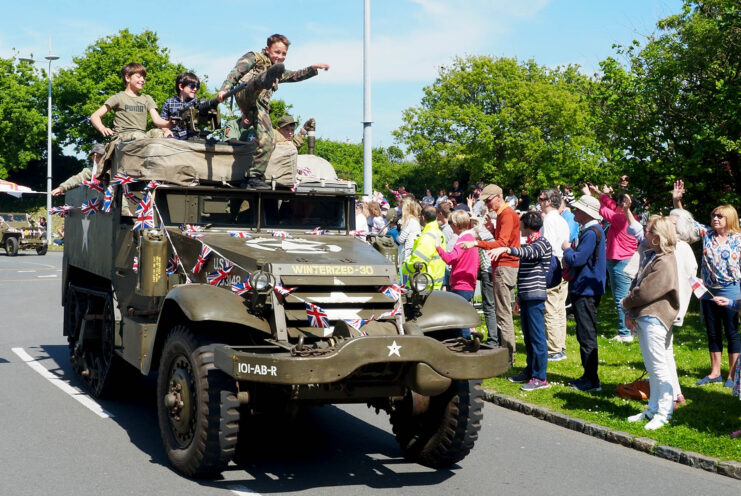 Children riding atop a military vehicle driving down a street lined with people