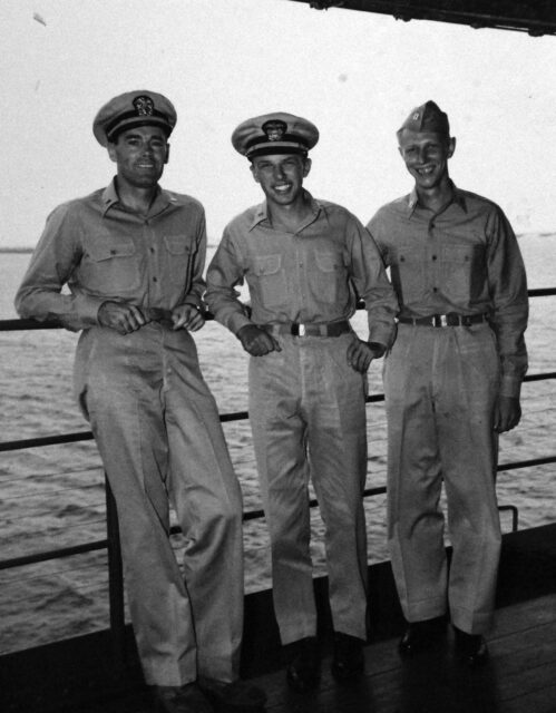 Black and white. Three men in US Navy uniforms lean on the railing of a ship and smile at the camera. Water in the background. 