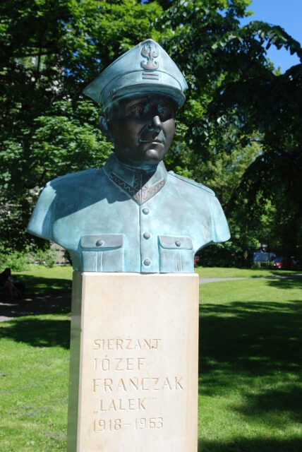 green bust of man in military uniform on top of white stone monument. The inscription on the white stone reads 'SIERZANT JÓZEF FRANCZAK LALEK 1918-1963'. Green grass and a tree in the background.