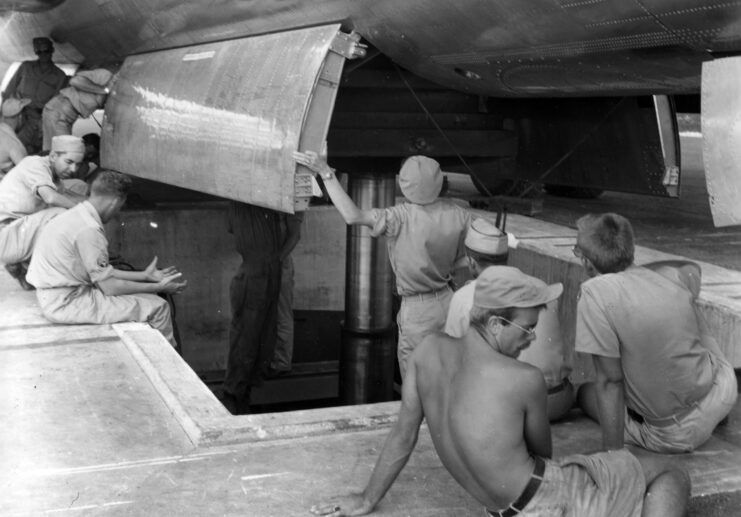 Black and white. Eight men in uniform shirts and pants and one man without a shirt sit or stand around a bomb pit in the tarmac and look up into the open bomb bay of a plane. 