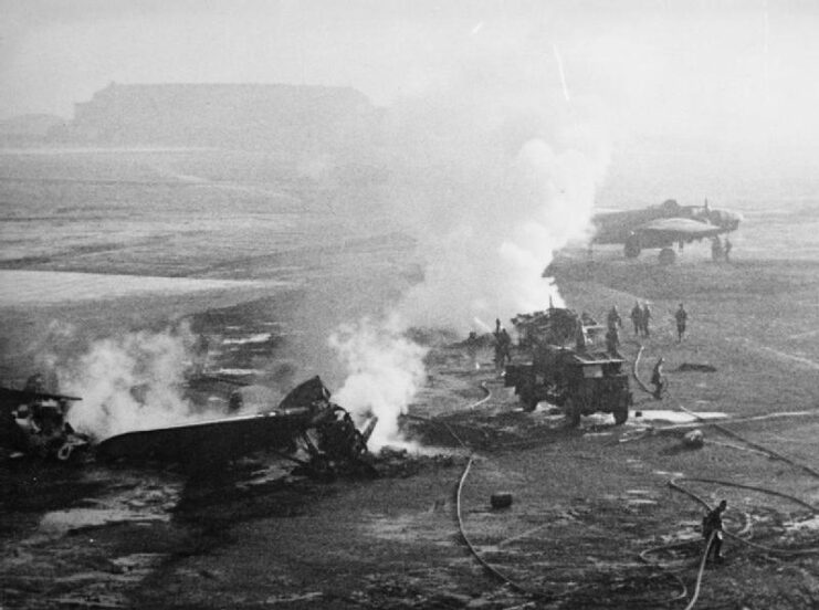 Aerial black and white image of two aircrafts with smoke billowing out of them on the ground. A vehicle approaches one aircraft and people holding hoses can be seen around the vehicles. 