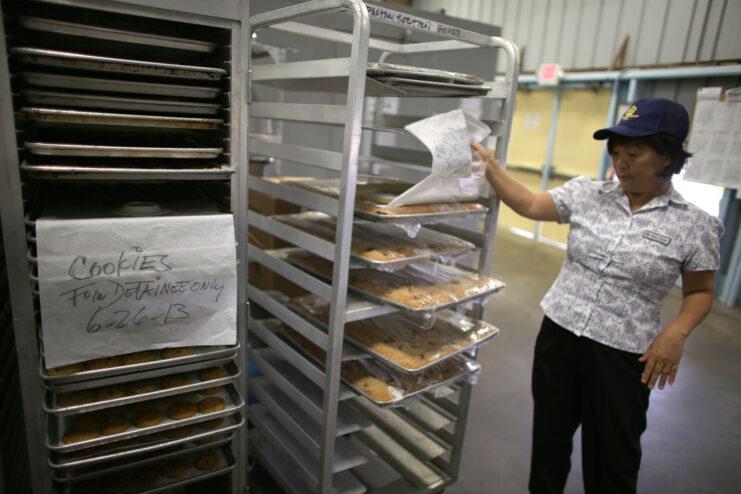 Woman in grey shirt and black hat pulls paper off of large silver tray of food that sits on a shelf of trays with baked goods. Paper attacked to shelves of trays reads 'COOKIES FOR DETAINEE ONLY.'