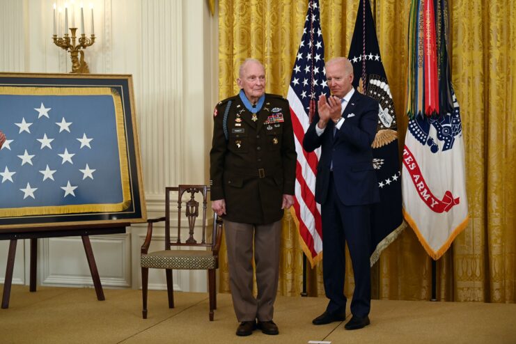 Man in black military uniform stands beside a man in a suit who claps as he wears a medal with a blue ribbon around his neck. A framed image of a ble background with 13 visible stars is to the left of both men and the flag of the United States of America and the American Military are behind them. 