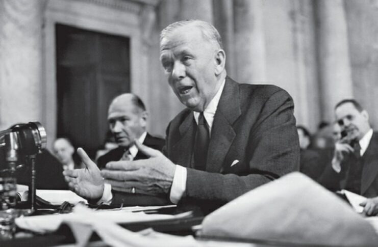 Man in suit speaking while sitting behind a desk. Two men can be seen behind him. Black and white. 