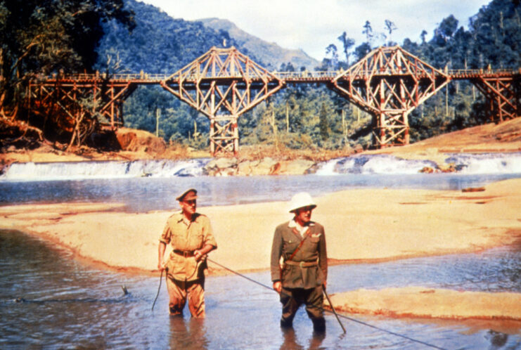 Two men in military uniforms satnd in knee-high water holding a rope with a bridge behind them. 