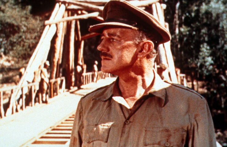 Alec Guinness looking off to the left, dressed in military uniform. Bridge can be seen behind him. 