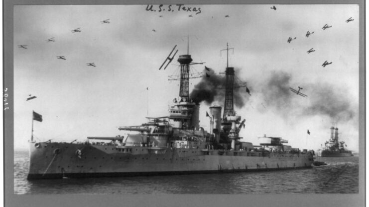 USS Texas (BB-35) at sea with another ship, while aircraft fly overhead