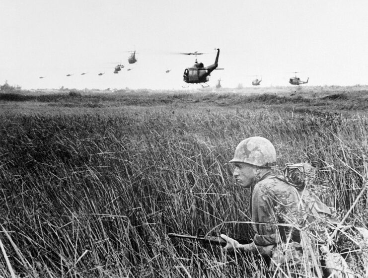 Helicopters flying away from a US infantryman crouching in tall grass