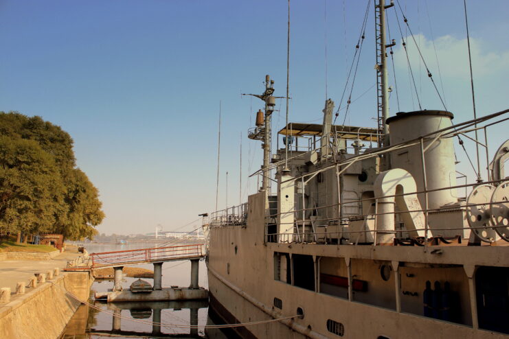 Left side of USS Pueblo docked close to the shore with a ramp. Trees on shore and blue sky. 