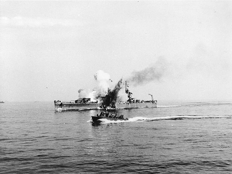 Small boat transiting past the USS Savannah (CL-42), which has smoke billowing from it