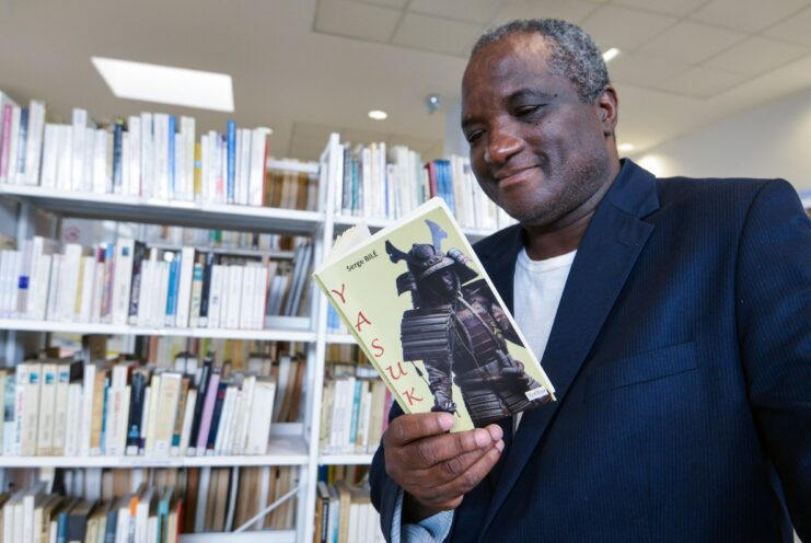 Serge Bile reading a copy of his book 'Yasuke' in a library. 