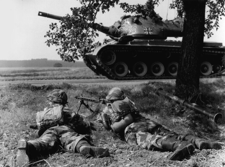 Two soldiers lying on the ground as an M47 Patton drives by