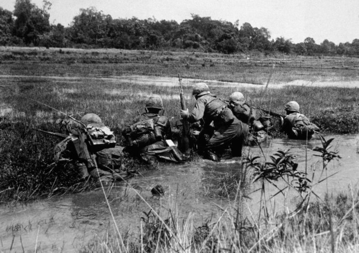 Five American soldiers aiming their weapons while crouching in a small waterway