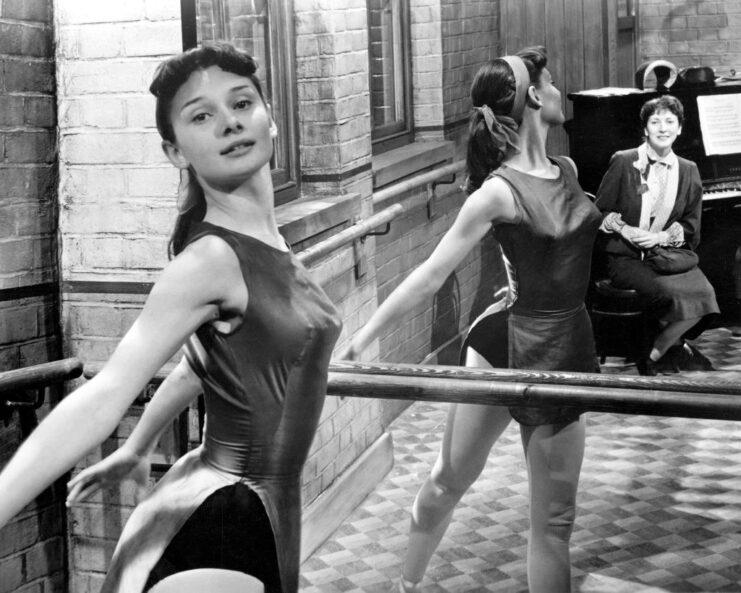 Audrey Hepburn standing at a ballet bar, while a woman looks at her