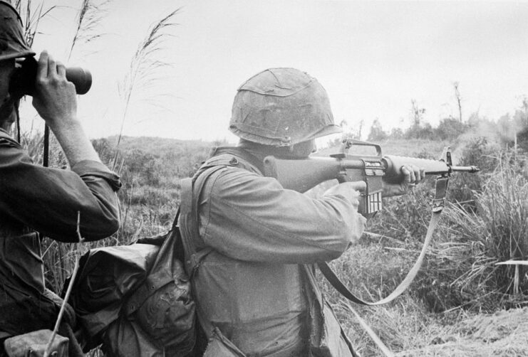 US Marine firing an M16 rifle, while another looks into the distance through binoculars