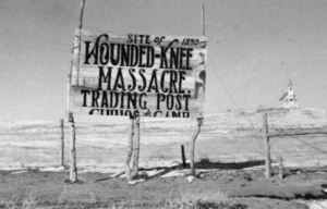 Sign in the middle of a field, with "SITE OF 1890 WOUNDED KNEE MASSACRE / TRADING POST" written on it