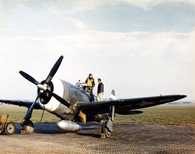 A P-47 Thunderbolt on an airfield with two men speaking on it. 