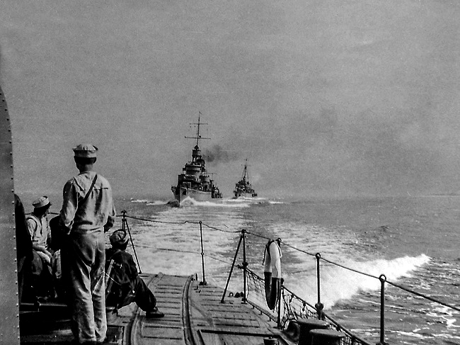 Sailors standing aboard the ORP Błyskawica, with the ORP Burza and Grom transiting in the distance