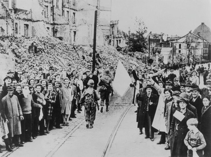 Crowd cheering on members of the Seventh US Army as they walk down a street surrounded by destruction