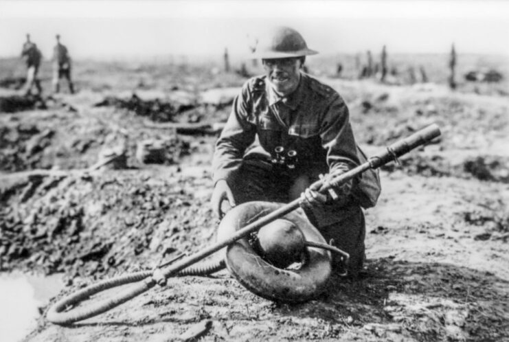 Australian soldier crouching while holding a captured German flamethrower in the middle of a battlefield